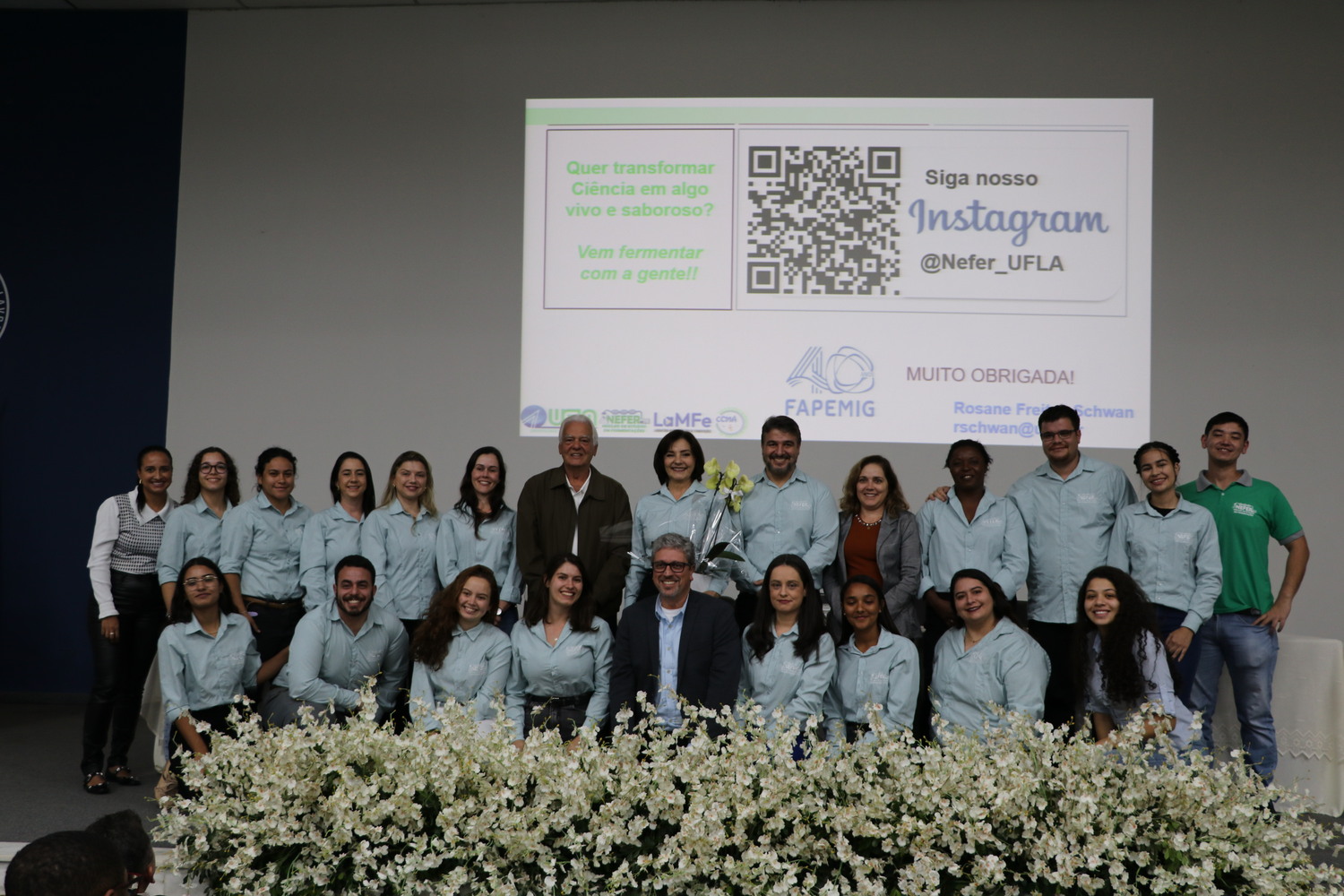 Na imagem, estão cerca de 30 pessoas sob um palco. Na frente delas um arranjo de flores. Eles estão de camisa azul dizendo que são do Nefer. O Núcleo de Estudos especializado em fermentação da Ufla;